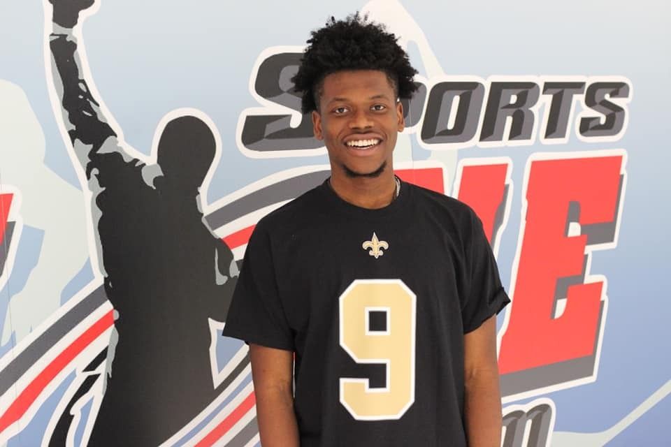 Smiling man in a black Saints jersey with the number 9, stands in front of a sports background.