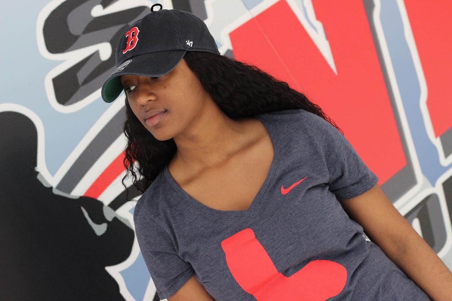 Woman wearing a Red Sox hat and shirt, looking to the side. Standing in front of a graffiti-style background.