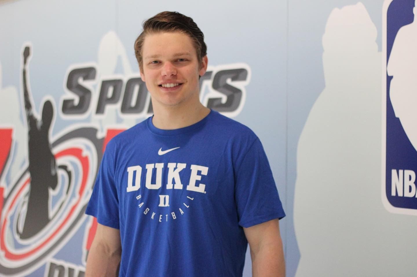 Man in blue Duke basketball shirt smiles, standing in front of a sports-themed background.