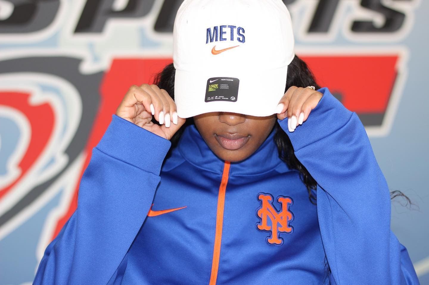 Woman wearing a white Mets hat and blue Mets jacket, adjusting the hat, in front of a blue background.