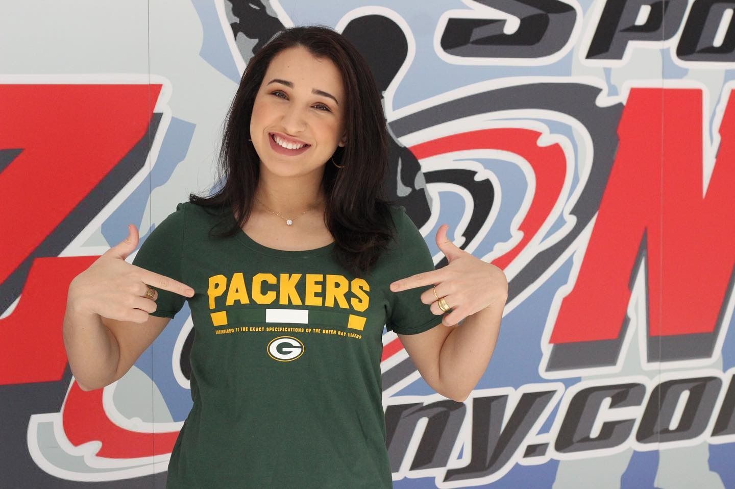 Woman in Packers shirt points at it, smiling; standing in front of a sports-themed backdrop.