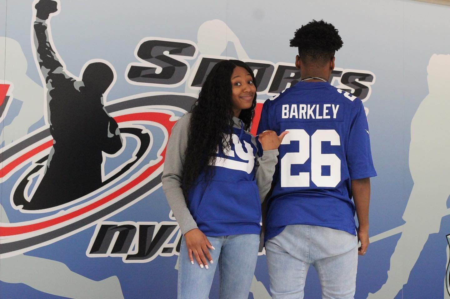 Two people wearing blue Barkley #26 Giants jerseys pose in front of a sports-themed wall.