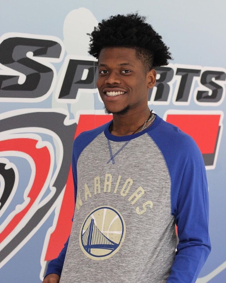Man with dark hair smiling, wearing a blue and gray Golden State Warriors sweatshirt, standing in front of a sports logo.