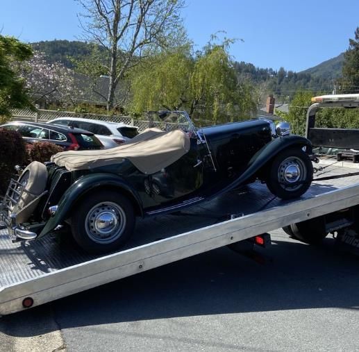 Dark green classic car on a tow truck, being transported on a sunny day.