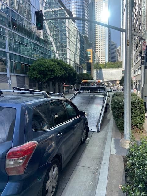 A blue car being loaded onto a flatbed tow truck on a city street. Green traffic light and buildings visible.