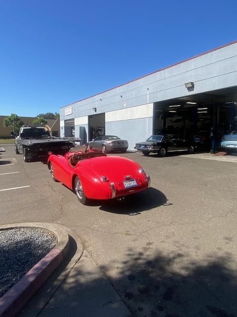Red classic car parked in front of a garage; other vehicles visible. Sunny day.