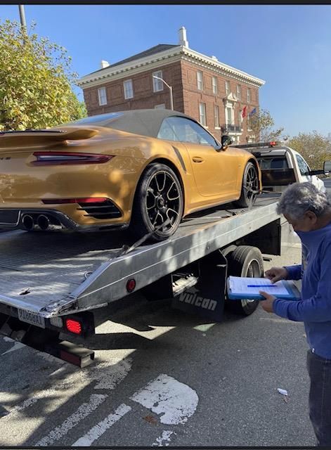 Gold convertible Porsche being loaded onto a tow truck by an older man in front of a brick building.