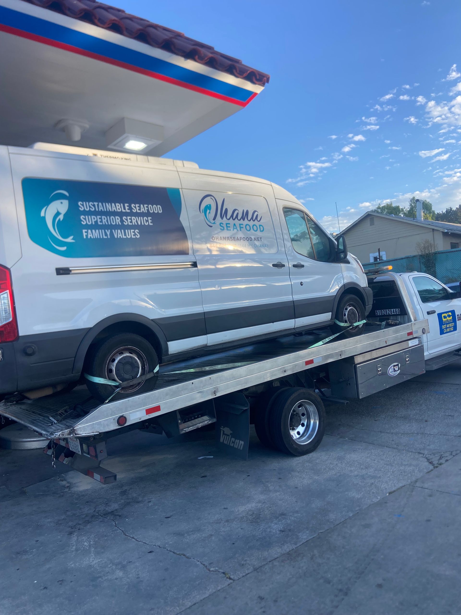 White service van being towed on a flatbed tow truck at a gas station.