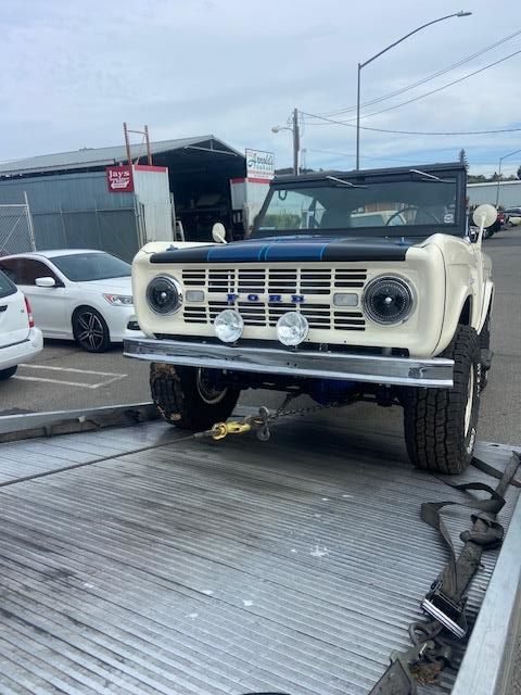 White and black vintage Ford Bronco on a tow truck, front view. Blue racing stripes on the hood.