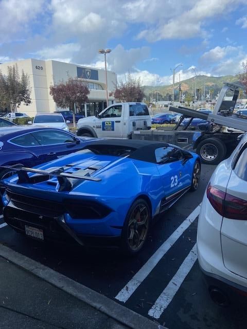 Blue Lamborghini Huracan convertible parked outside, next to a white SUV and a tow truck, under a cloudy sky.