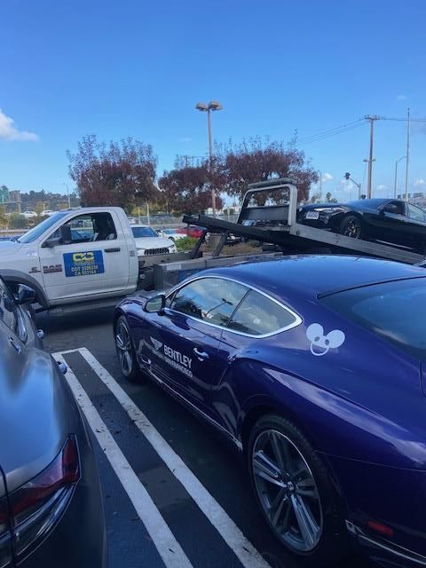 Blue Bentley being towed in parking lot on a sunny day, tow truck nearby.