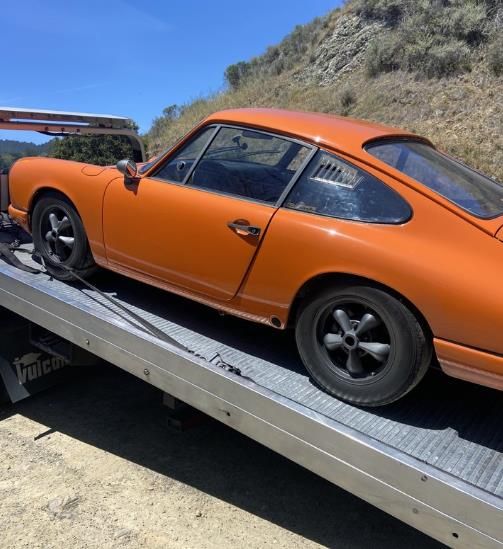 Orange vintage Porsche on a flatbed tow truck. Mountain backdrop on a sunny day.