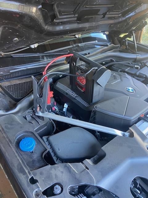 A car engine with a jump starter connected. Black engine, open hood, red and black cables.