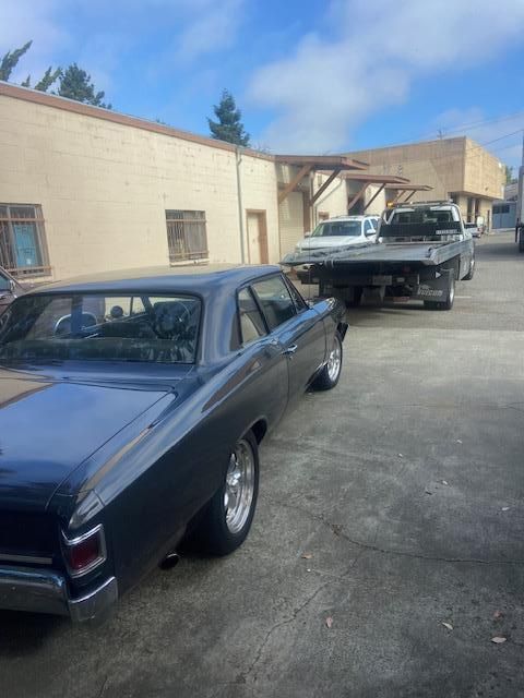 Dark blue classic car in front of a white building with a tow truck in the background.
