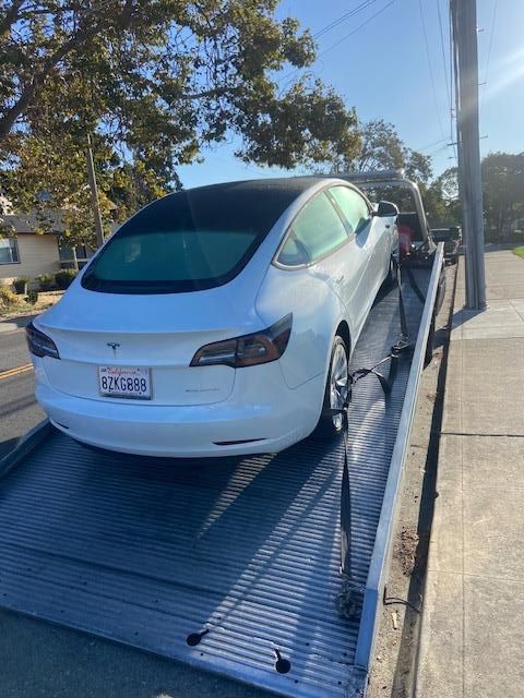 White Tesla Model 3 on a tow truck, likely being transported. Sunny outdoor setting.