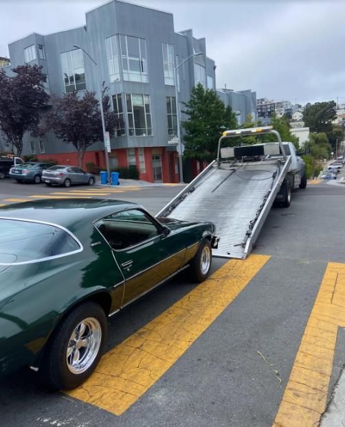 Green classic car being loaded onto a tow truck on a city street with a modern building in the background.