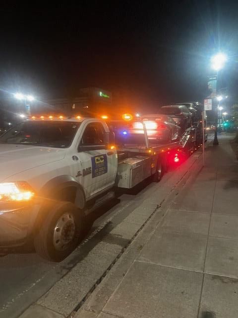A white tow truck with flashing lights loading a car onto its flatbed at night.