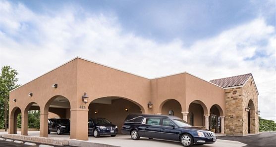 Tan stucco building with arched carport, several parked cars, and stone accents under a cloudy sky