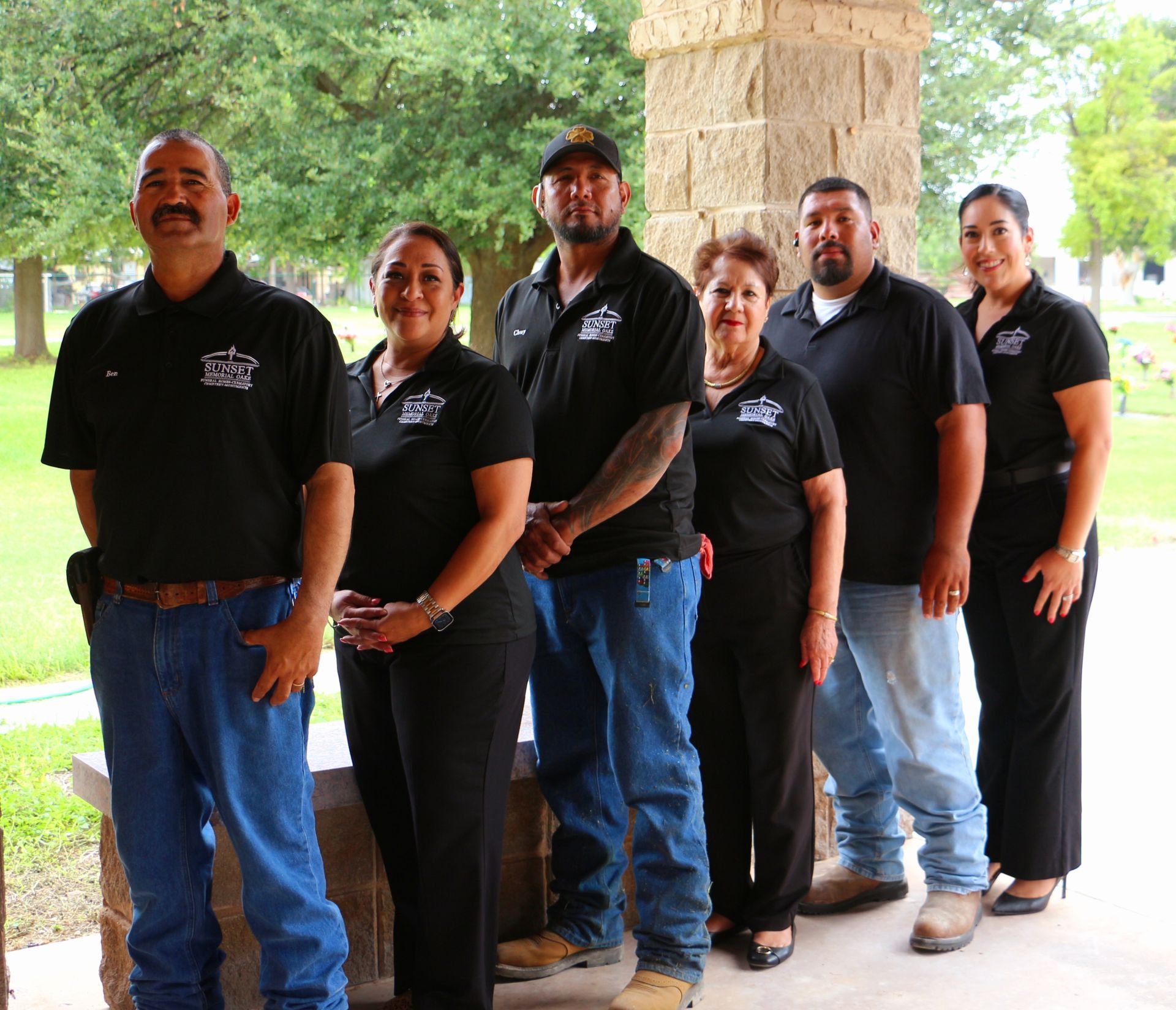 Six people in black shirts standing outdoors by a stone pillar, with trees in the background.