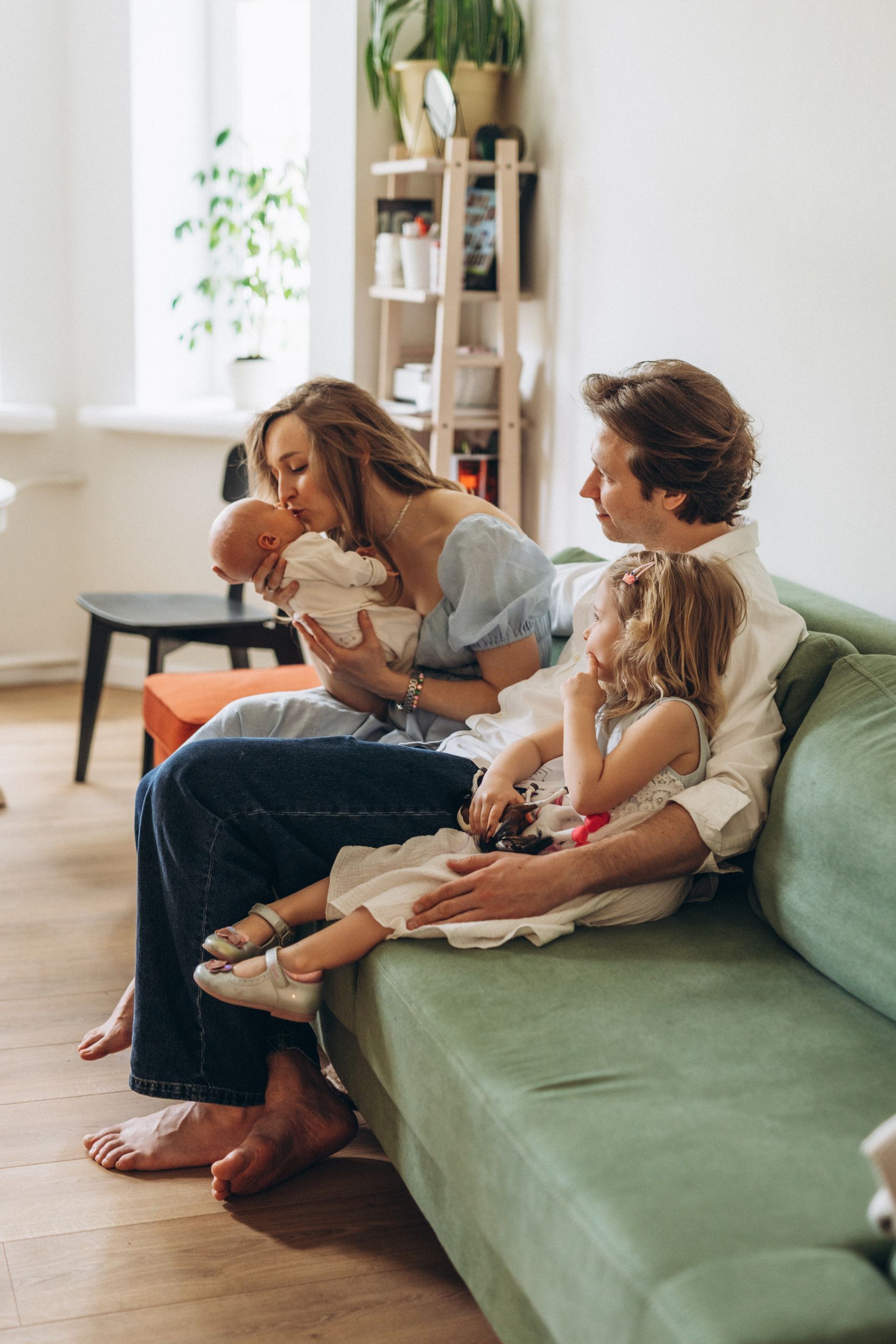 Woman on green sofa with two children, one holding a stuffed toy, in a bright living room.