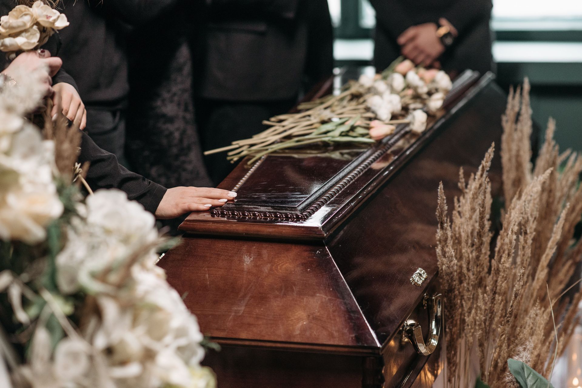 A casket at a funeral surrounded by white flowers and mourners in dark clothing