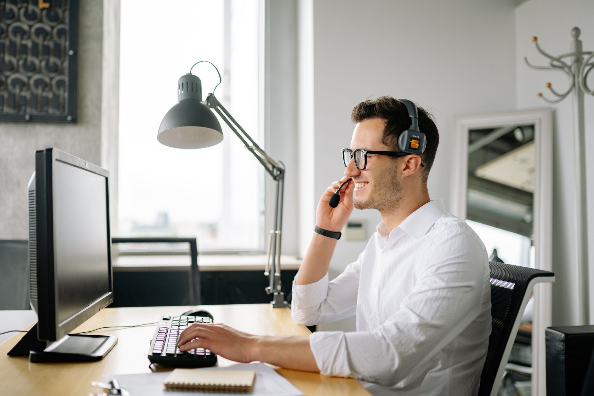 A man is sitting at a desk in front of a computer and talking on a cell phone.