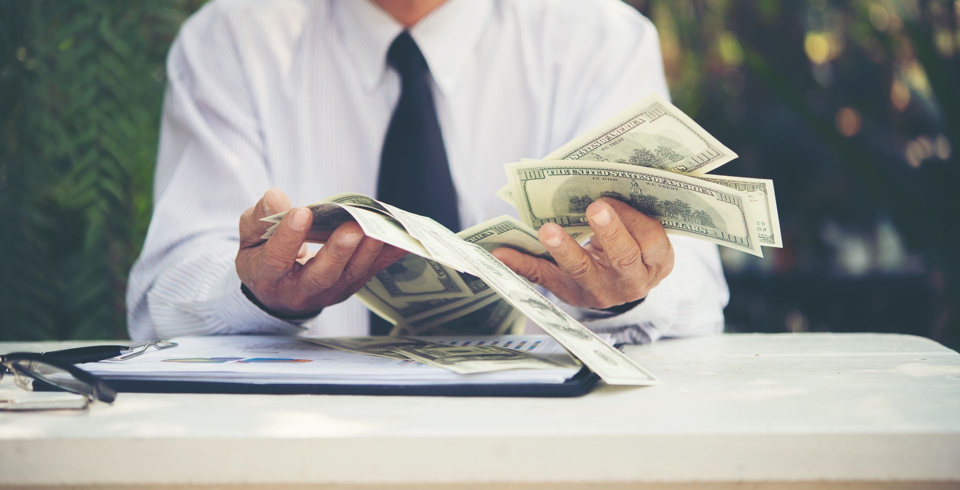 Man in collared shirt and tie with hundred dollar bills in his hands sitting at a desk