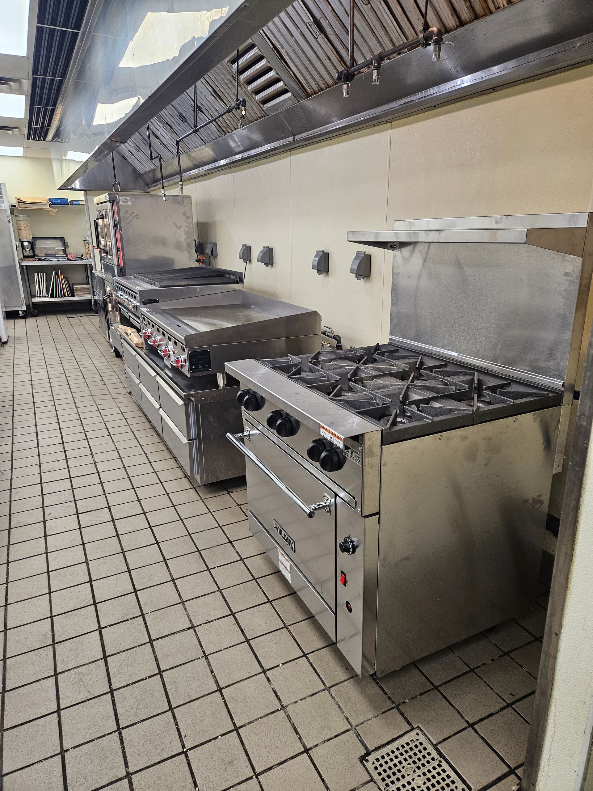 Commercial kitchen with stainless steel appliances under a ventilation hood. Tile floor.