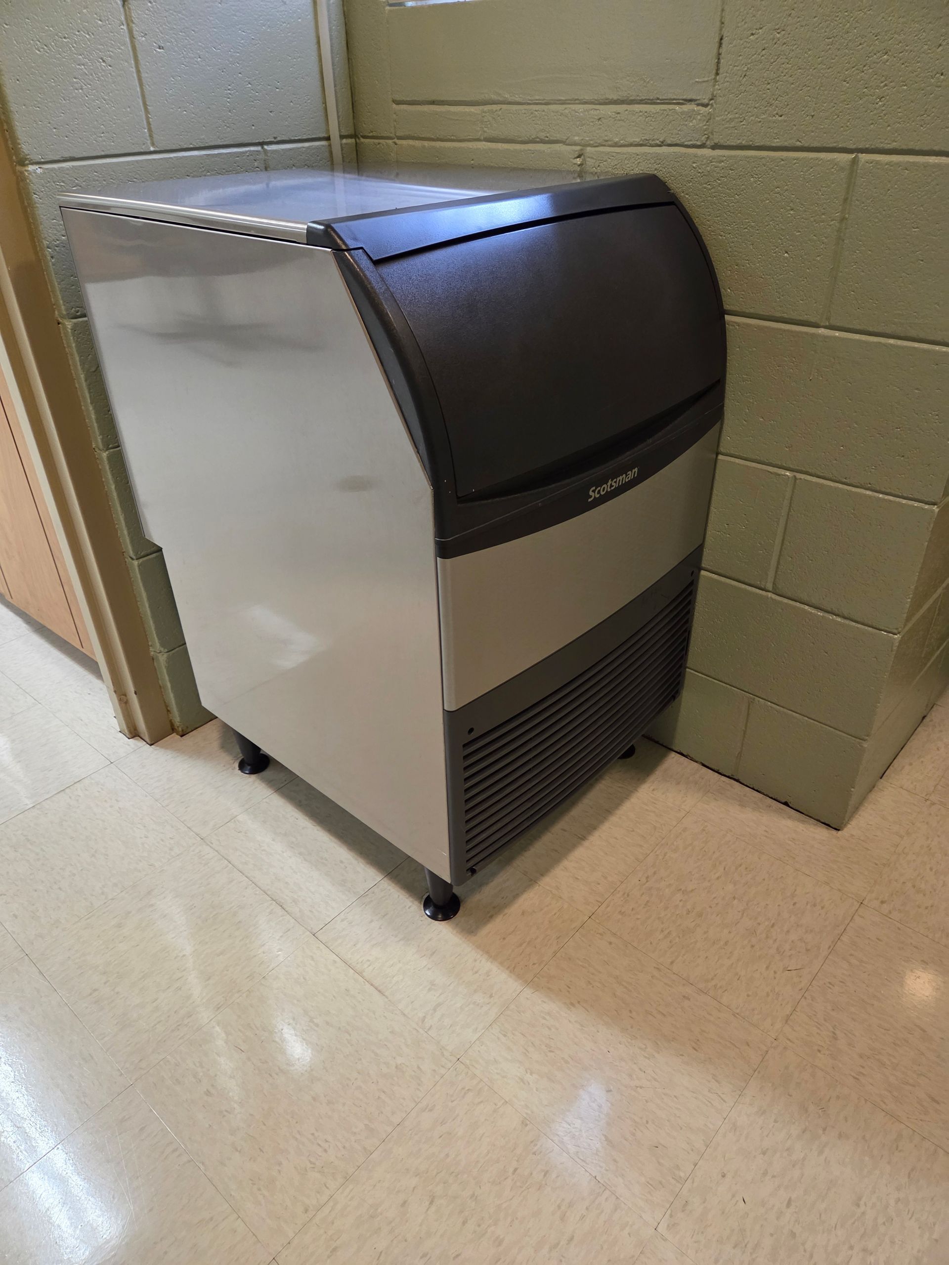 A silver and black ice machine on wheels sits next to a cinder block wall.