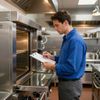 Man in blue shirt, inspecting commercial oven, writing on clipboard in kitchen.