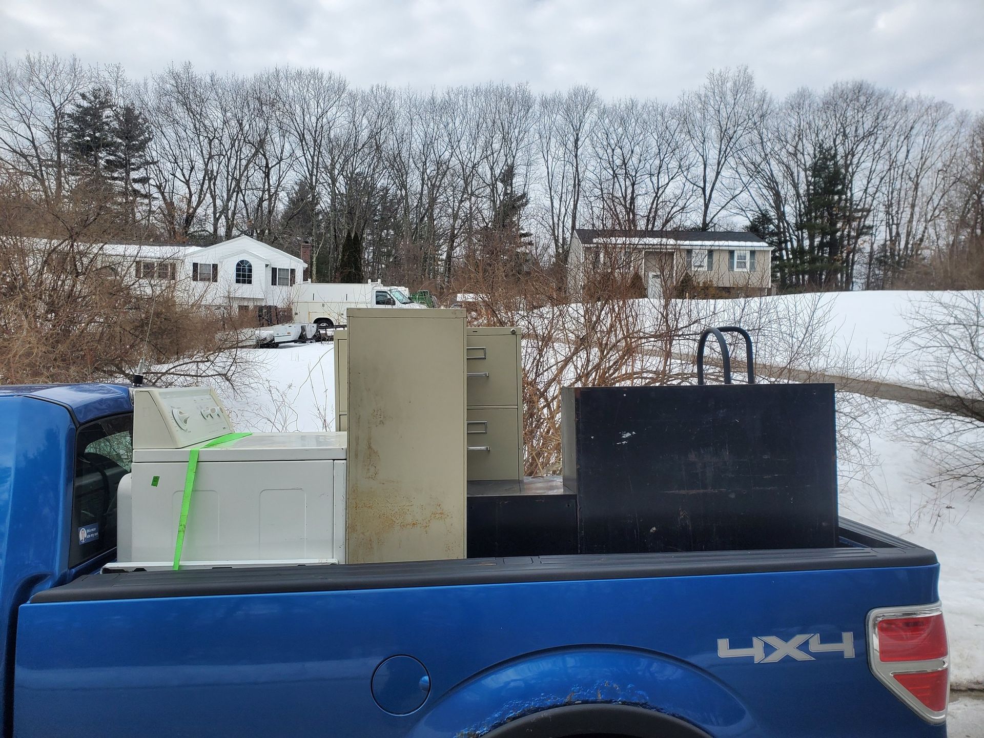 Blue truck bed loaded with a washing machine, filing cabinets, and a toolbox; snowy suburban background.