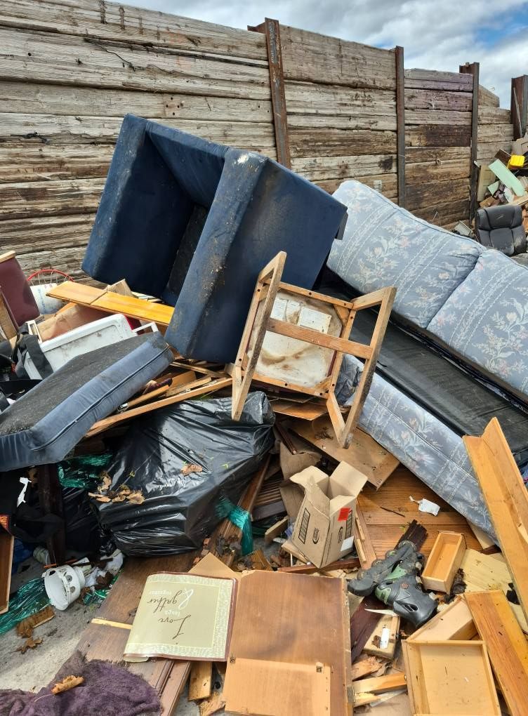 Pile of discarded furniture and trash against a wooden wall, including a blue couch, chair frame, and black trash bags.