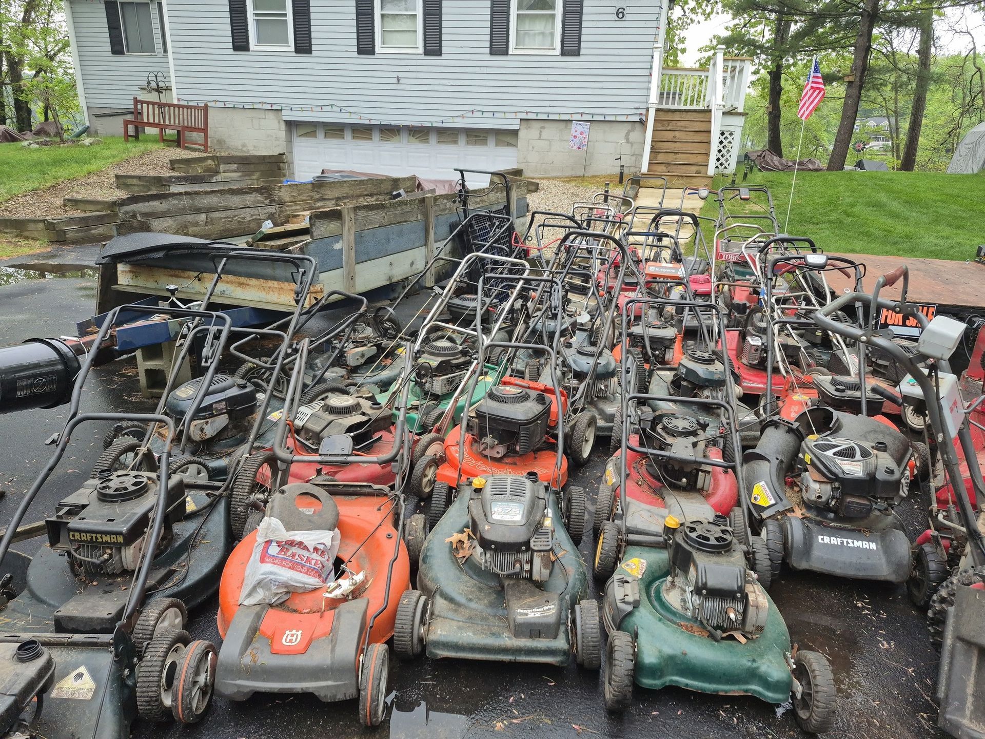 Many lawn mowers clustered together in a driveway; house in the background.