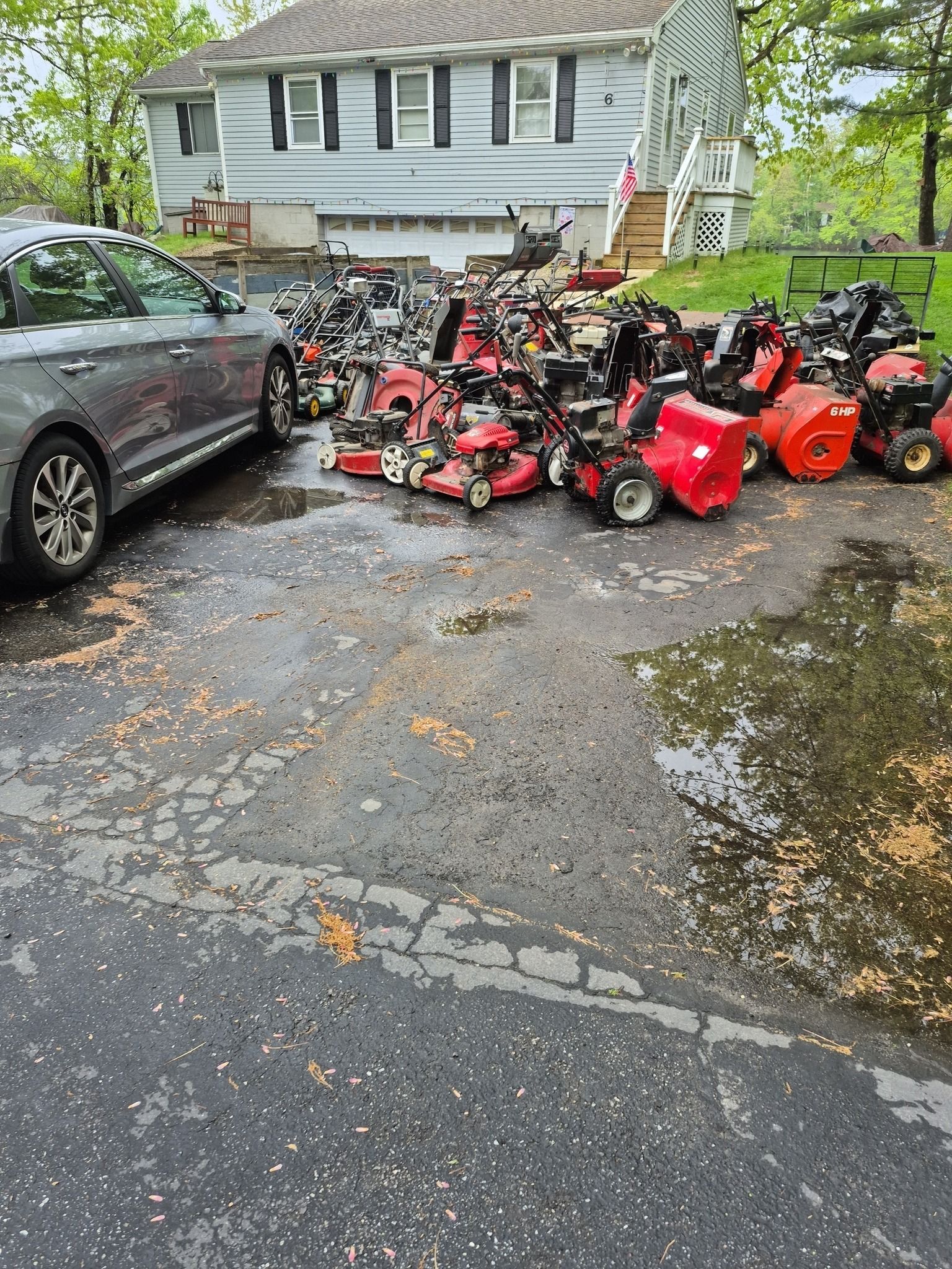 A large pile of lawn mowers and snow blowers on a wet driveway near a house and a car.
