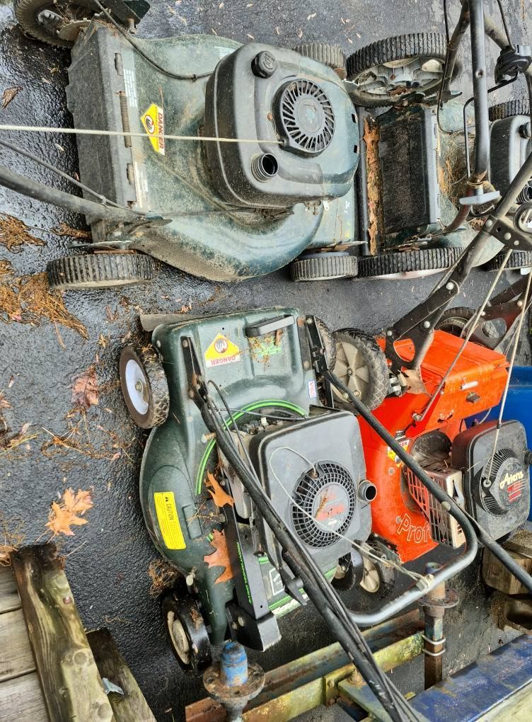 Three lawnmowers of varying sizes and colors are arranged outdoors on a black surface.