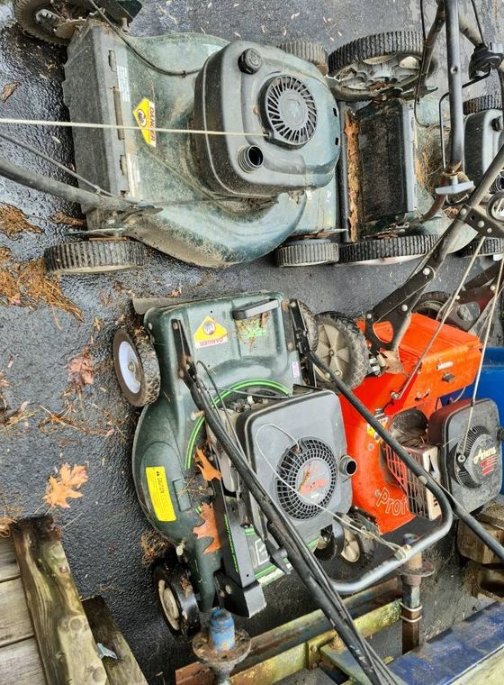Four lawn mowers, various colors, sit outdoors, possibly on a concrete surface.