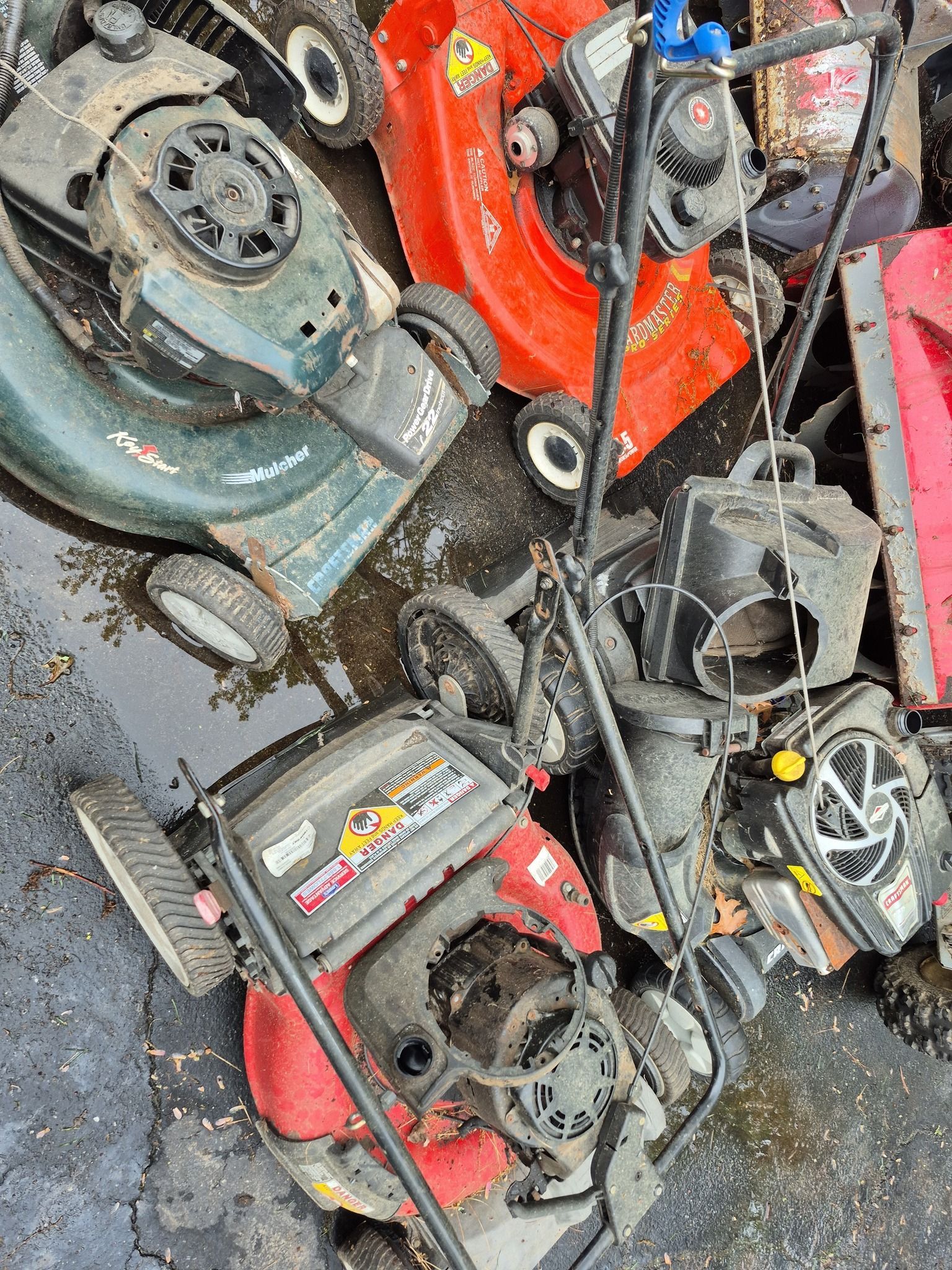 Pile of discarded lawnmowers in various colors: red, green, and gray. Outdoors, on a wet surface.