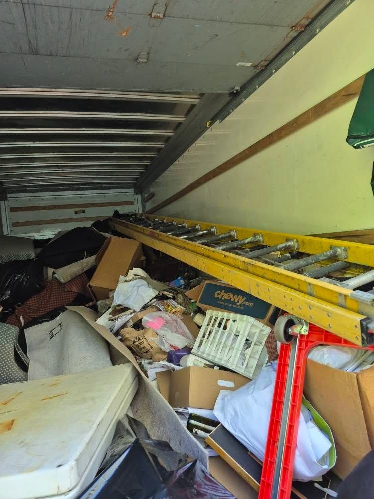 Inside a cluttered truck, a yellow ladder leans over debris, boxes, and a mattress.