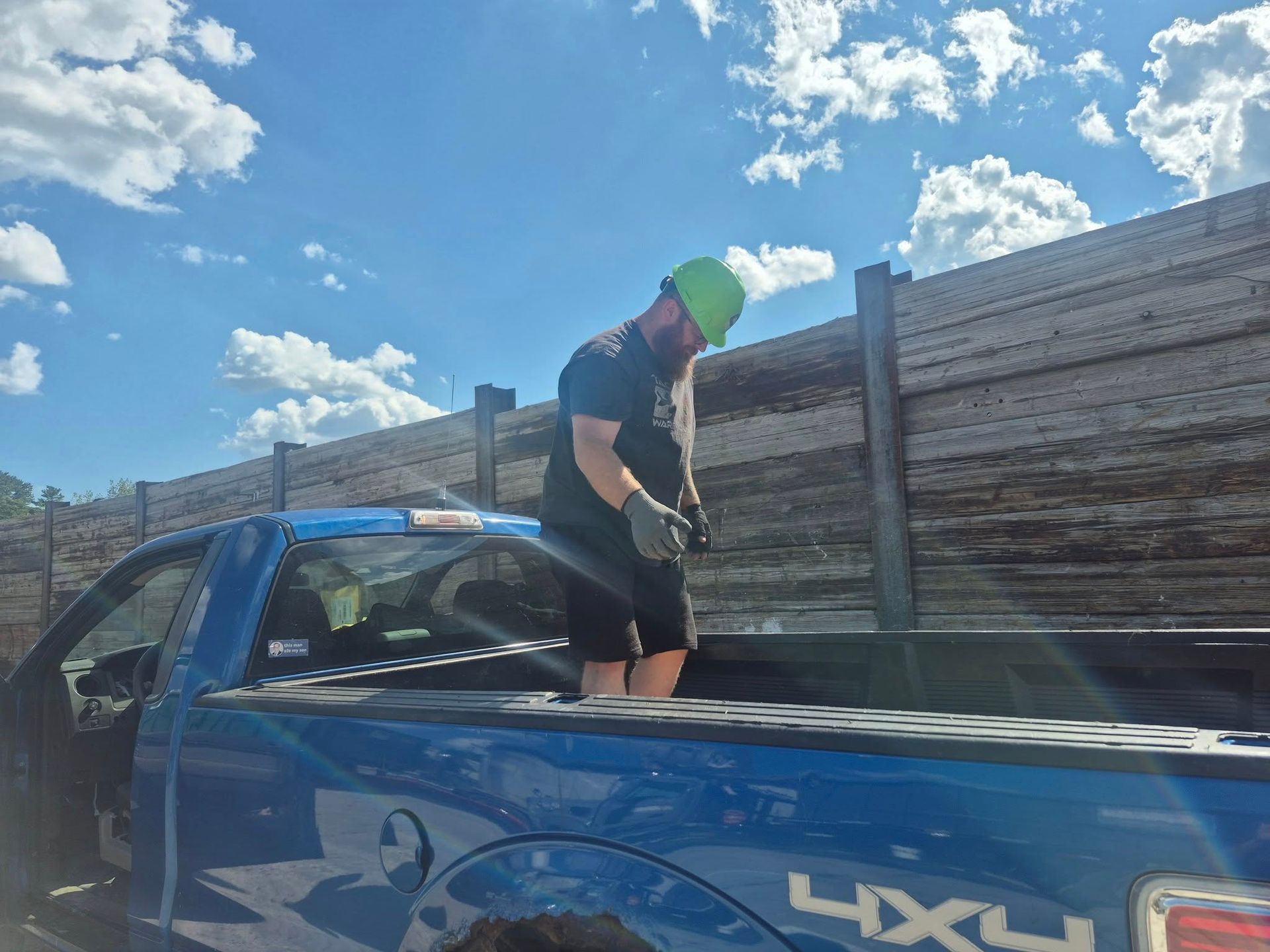 Man loading wood planks into the bed of a blue pickup truck on a sunny day, near a wooden fence.