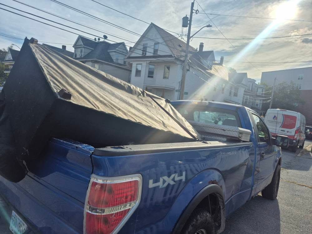 Blue pickup truck with a black mattress in the bed, parked on a street with houses.