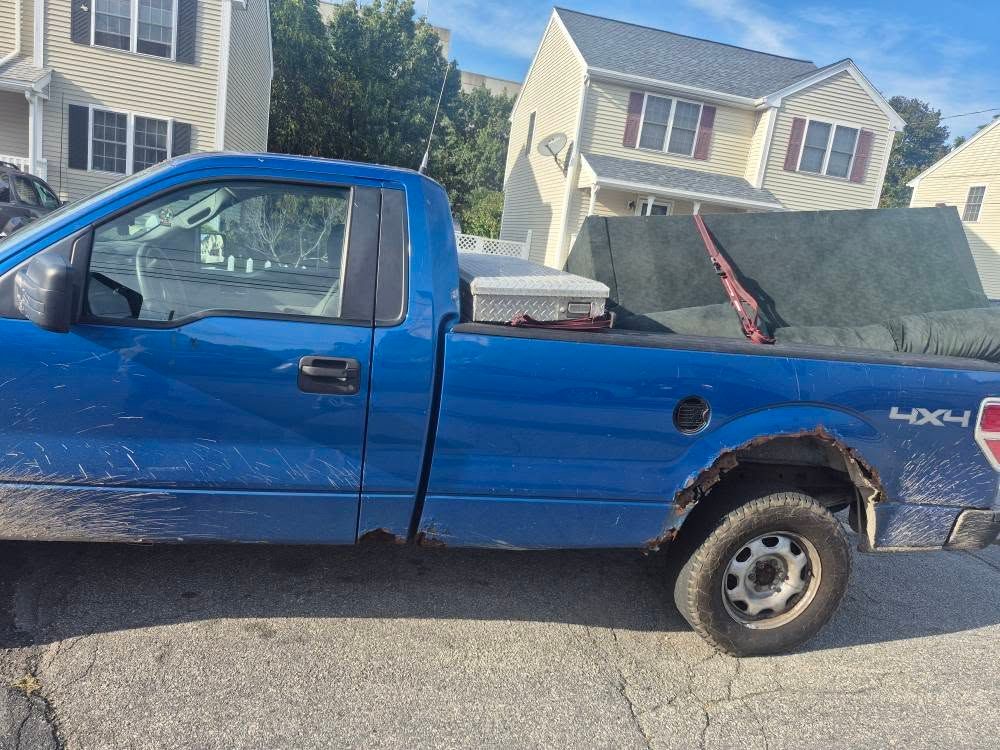 Blue pickup truck with a couch in the bed parked on the side of a road, next to a two-story house.