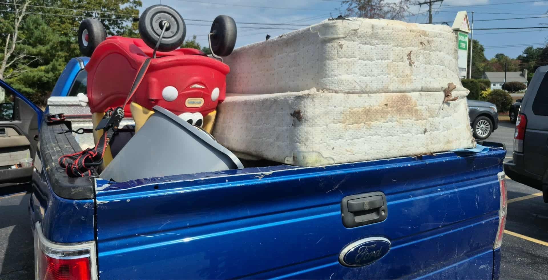 Blue pickup truck bed loaded with a child's toy, bucket, and stacked old foam cushions.