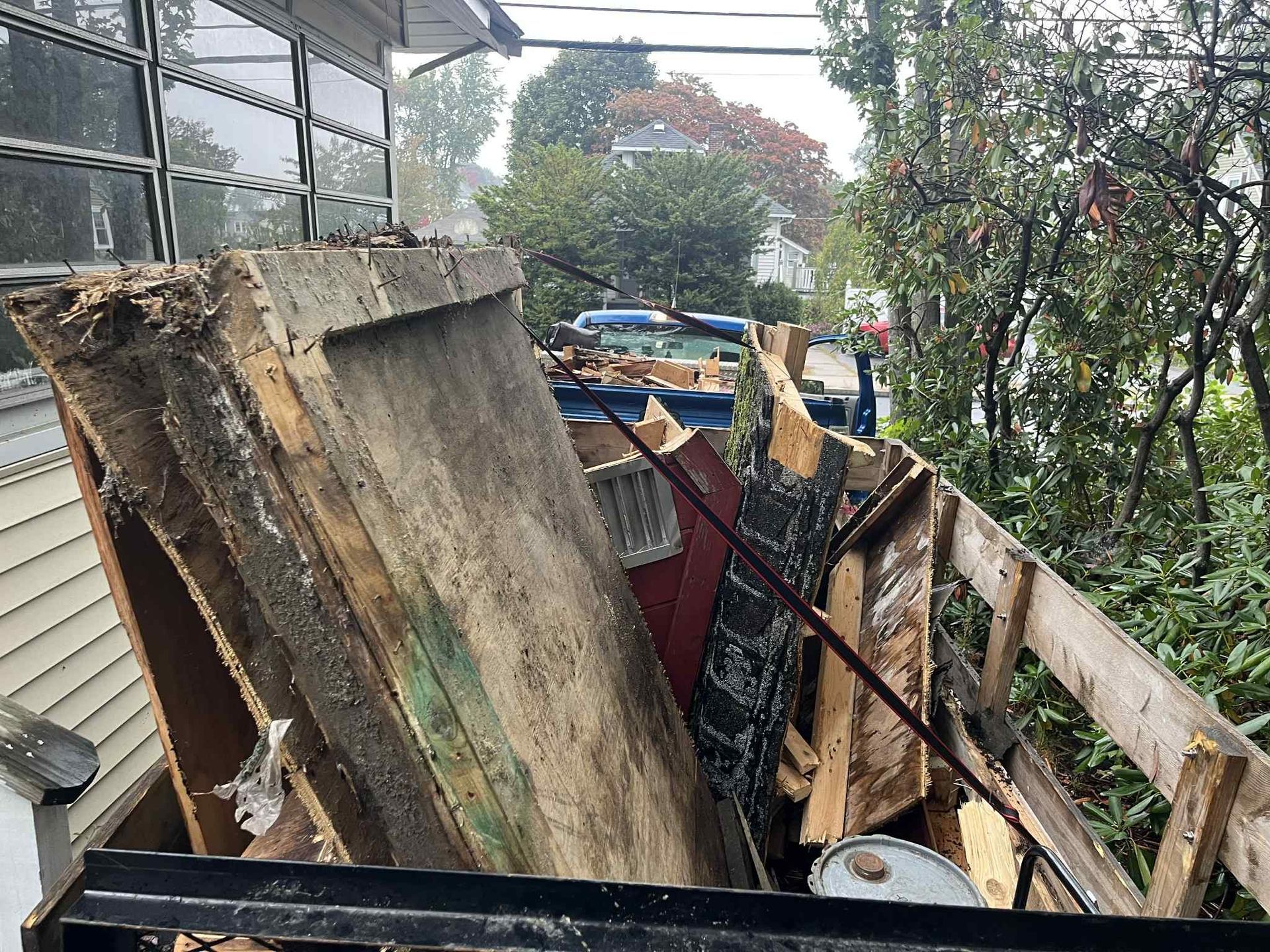 Debris-filled dumpster in front of a house; wood, metal, and paint cans, overcast day.