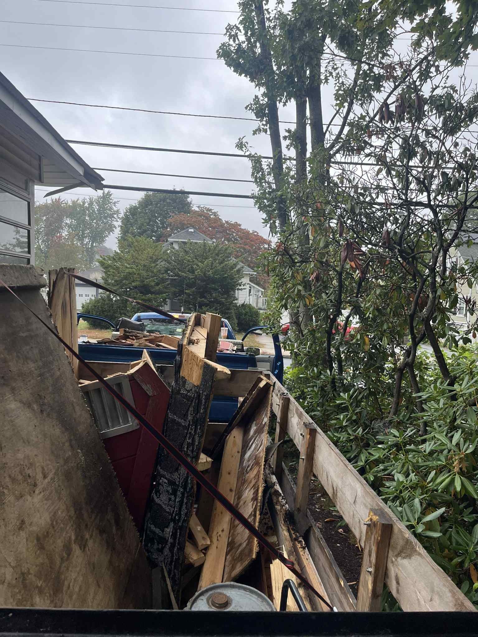 Pile of wood debris in a truck bed; trees and houses in the background under an overcast sky.