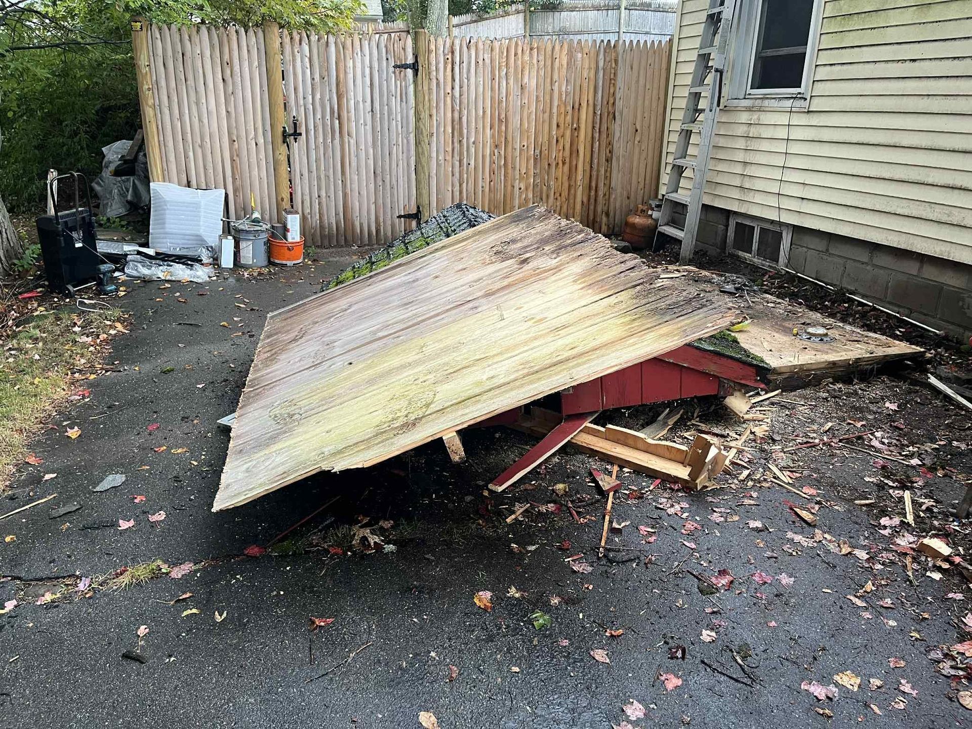 Fallen shed on dark asphalt next to a wooden fence and a yellow house. Debris scattered around.