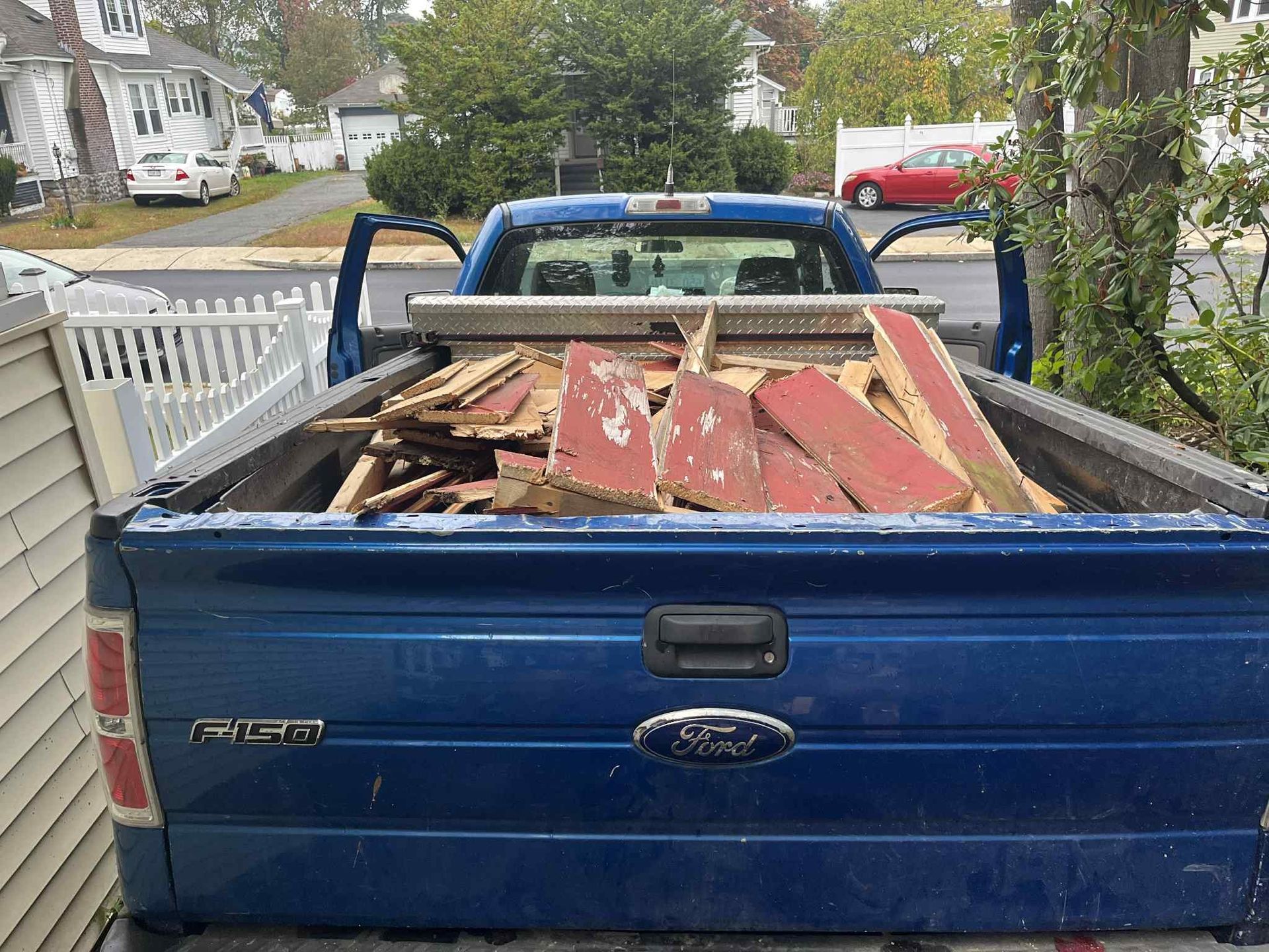 Blue pickup truck filled with old red and white wood debris, parked in front of a house with the doors open.
