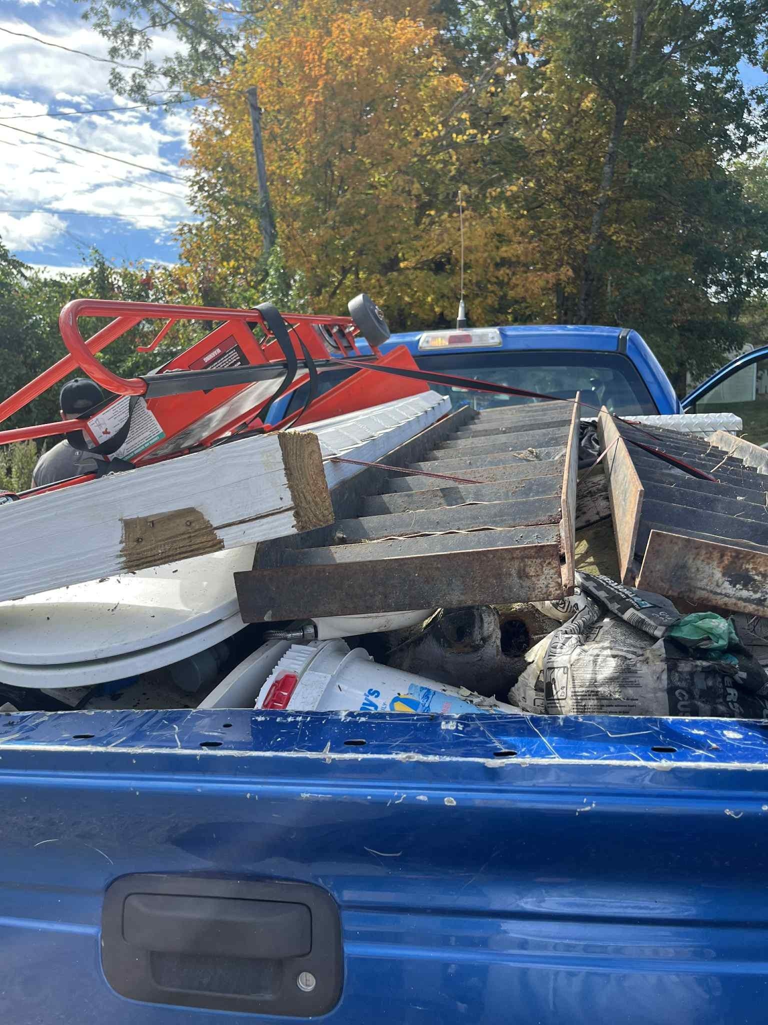 Blue truck bed filled with debris: white, red, and metal objects, set against fall foliage.