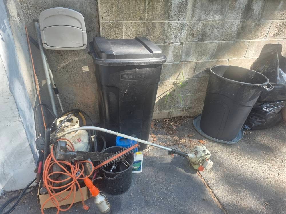 Equipment and trash containers leaning against a brick wall: weed wacker, hedge trimmer, trash cans, extension cord.