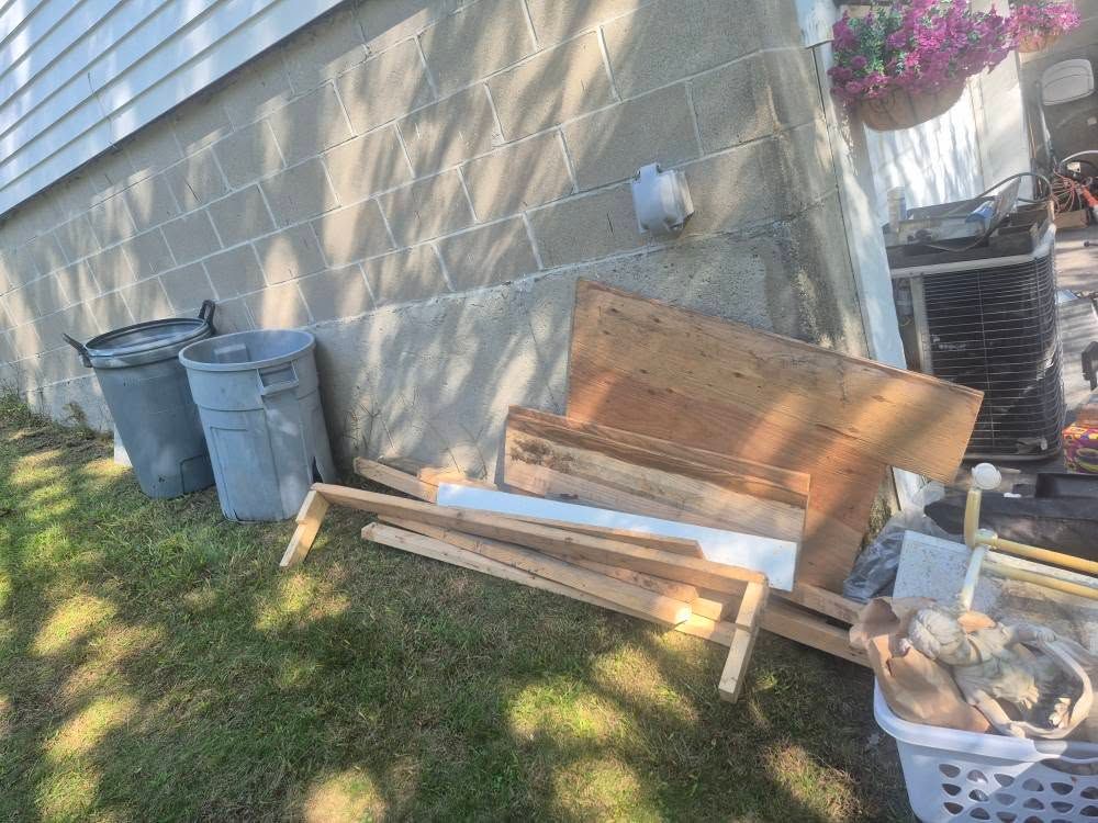 Two trash cans, lumber, and debris next to a concrete wall with a basket and air conditioning unit on the right.