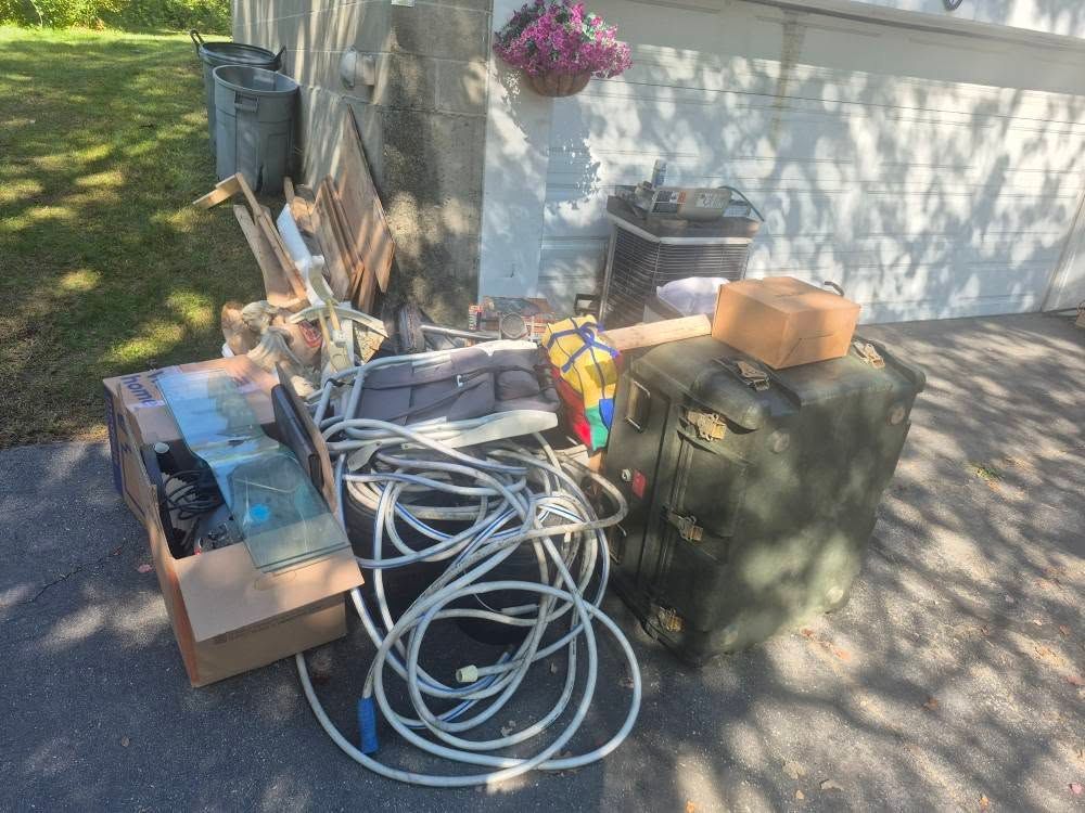 Pile of discarded items: cardboard box, hose, green bag, next to a wall with hanging flowers on a driveway.